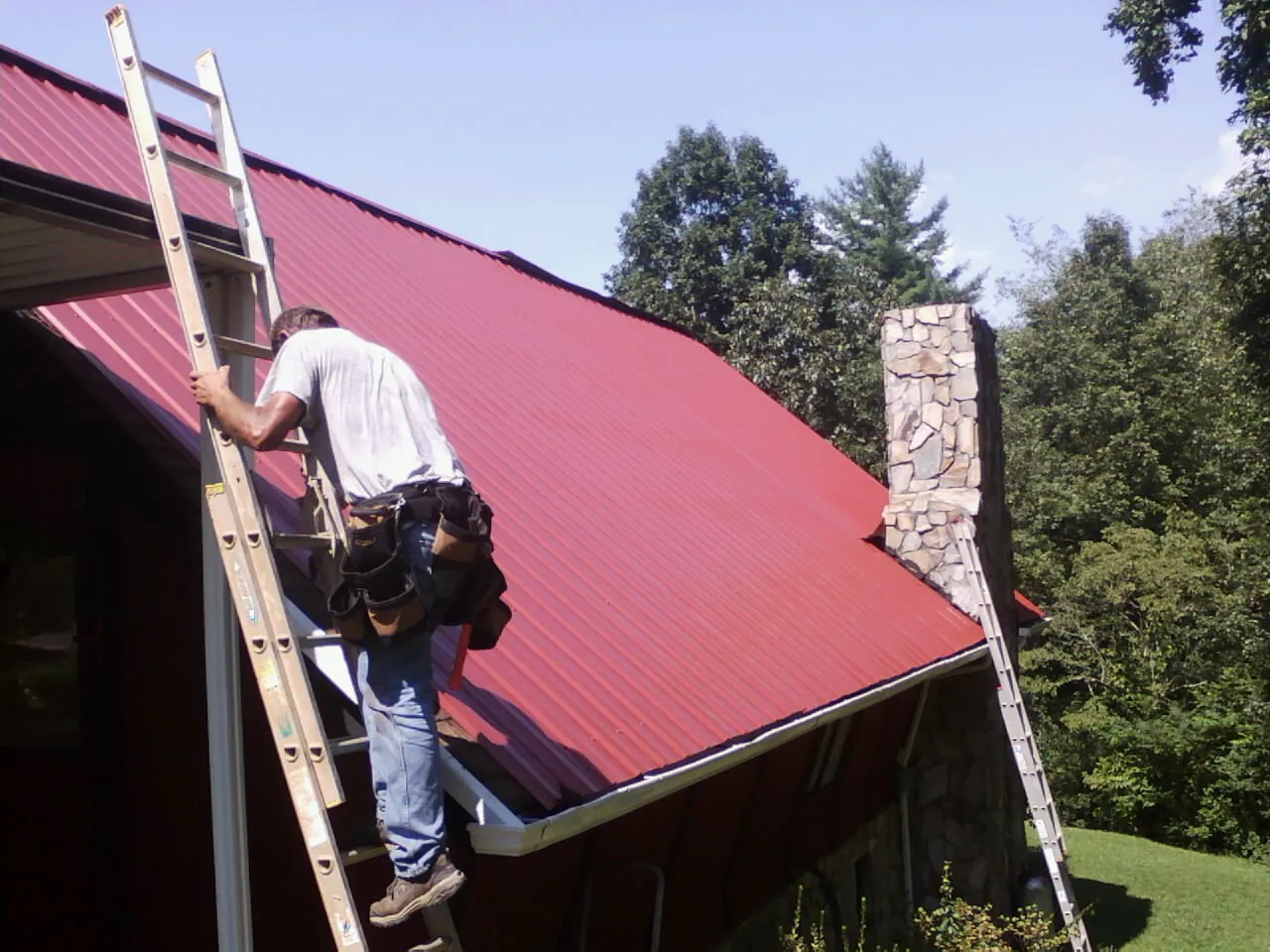 Roofer on a ladder beside a red metal roof and stone chimney — Soffit & Fascia Repair by Roofing Brothers in Leicester